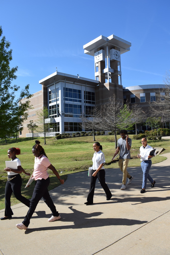Five people walk along a campus pathway outside a modern building with a clock tower. They carry notebooks and folders under a clear blue sky.