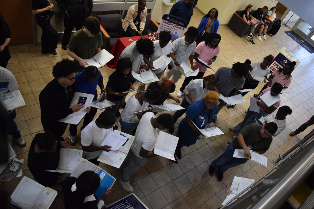 A group of people stand in a hallway holding clipboards and papers, completing forms. A table with a red cloth displays a sign about Pulaski Tech Tuition Match.