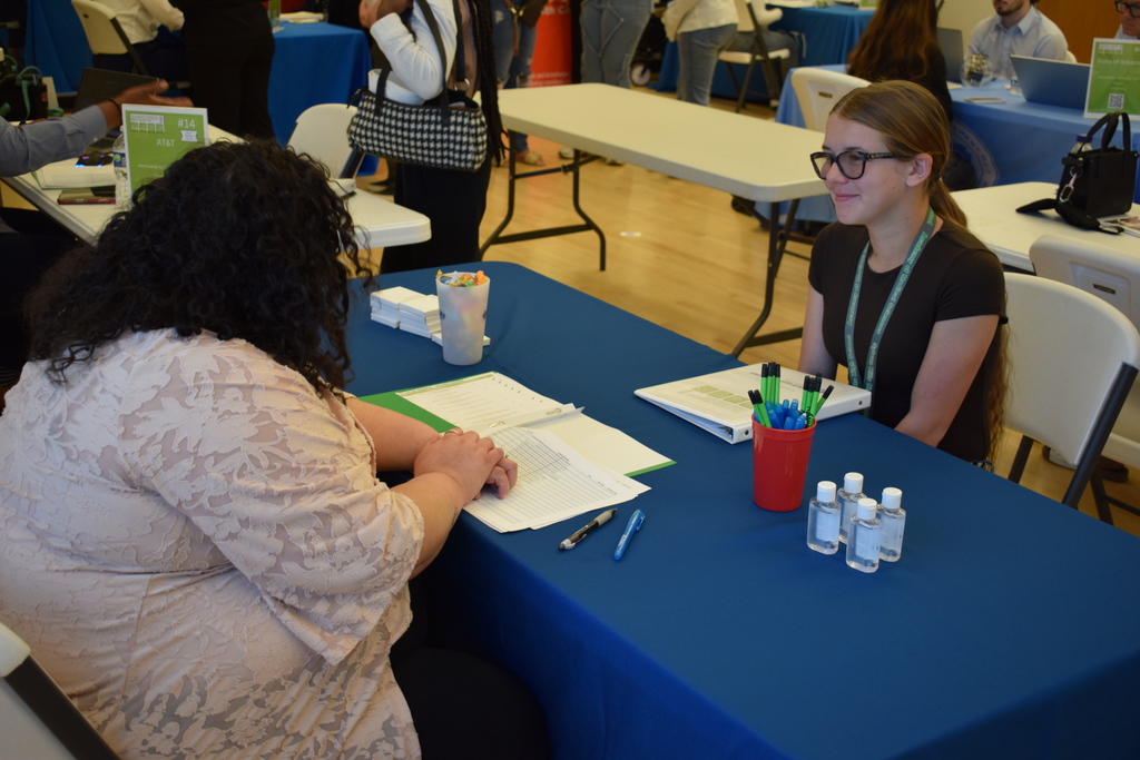 Two people sit across from each other at a table covered with a blue cloth, talking during a hiring event. Papers, pens, sanitizer, and a cup of candy are on the table. 