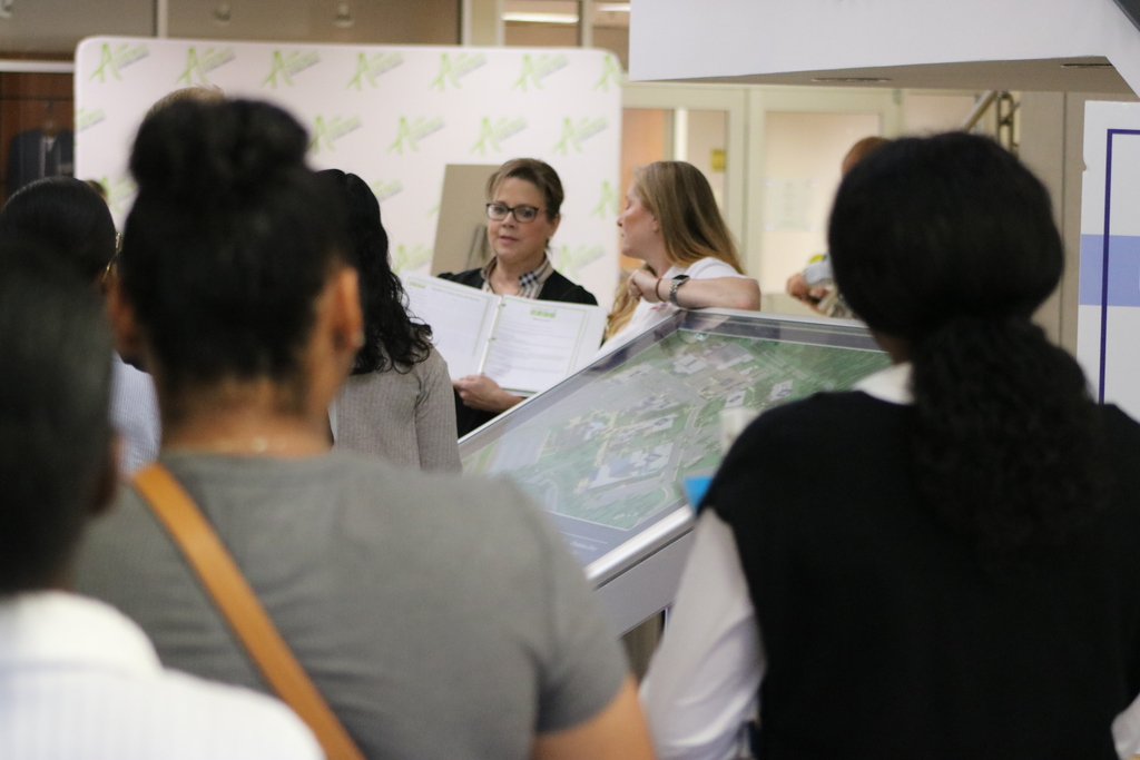 A group listens to a presenter holding an open binder while another person stands beside a large interactive map display. An Academies of Central Arkansas  backdrop is behind them.