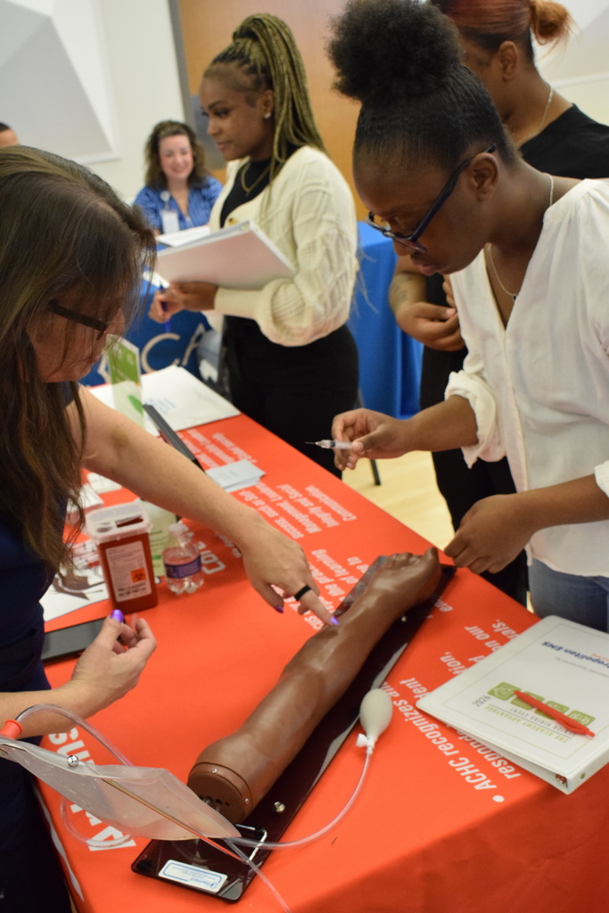 A student tries to stick a mannequin arm with a needle. She is being instructed by an adult from the other side of a table.