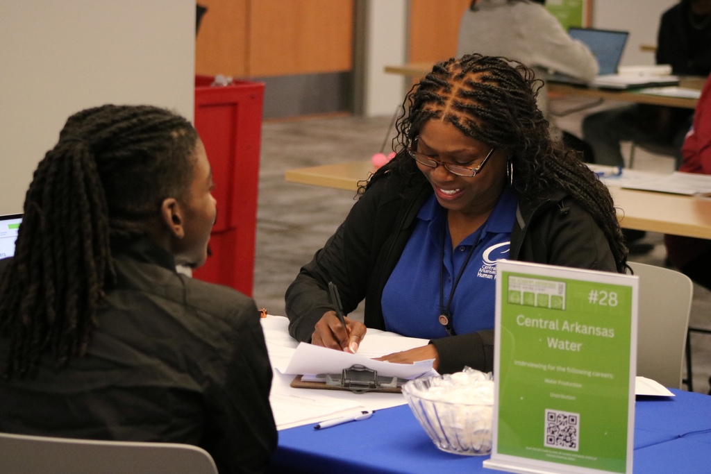 A representative from Central Arkansas Water speaks with a student across a table. A sign lists available careers in water production and distribution. Papers and a bowl of mints sit on the table.