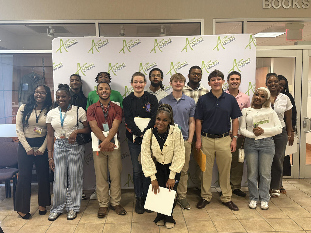 Fifteen students stand together in front of an “Academies of Central Arkansas” backdrop. They are dressed in business casual attire and holding folders or papers.
