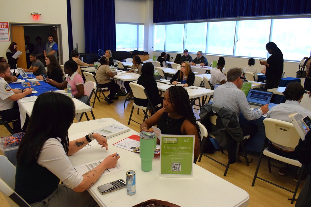 A large room filled with tables where students and employers sit across from each other conducting interviews. A sign on one table reads “#2 Arkansas Children’s.”