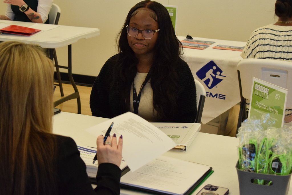 A representative from CARTI speaks with two students at a table covered with documents and a bottle of BodyArmor. A sign lists available careers such as nursing technicians and patient transporters.