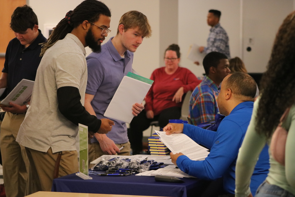 A group of people stand around a table at a hiring event. A representative hands a document to a student while others wait with folders and resumes. The table is covered with papers and promotional items.