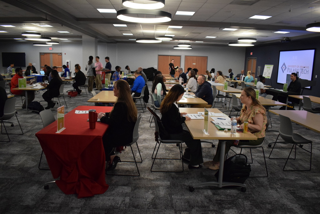 A wide view of a career event with rows of tables where pairs of people are engaged in interviews. A large screen in the background displays the University of Arkansas–Pulaski Tech logo.
