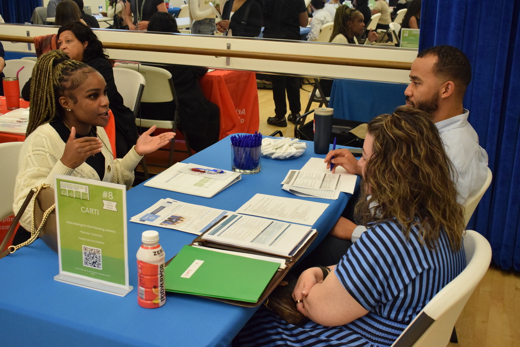 A representative from CARTI speaks with two students at a table covered with documents and a bottle of BodyArmor. A sign lists available careers such as nursing technicians and patient transporters.