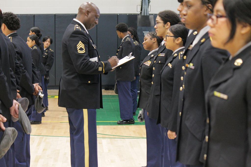 A U.S. Army JROTC instructor holding a clipboard while inspecting a line of Mills High School cadets, who are standing at attention in their dress uniforms.