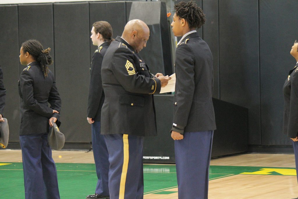 A U.S. Army JROTC instructor holding a clipboard while inspecting a line of Mills High School cadets, who are standing at attention in their dress uniforms.