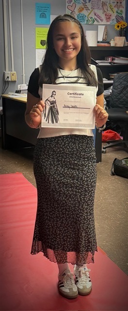 A student stands indoors on a red carpet holding a certificate. They wear a black top with a white collar, a long black floral skirt, and white sneakers.
