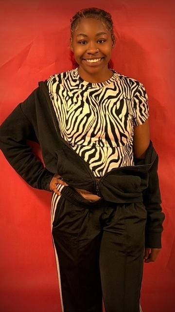 A student stands in front of a red backdrop wearing a zebra‑print shirt, a black jacket draped off one shoulder, and black pants with white side stripes. One hand rests on the hip in a runway‑style pose.