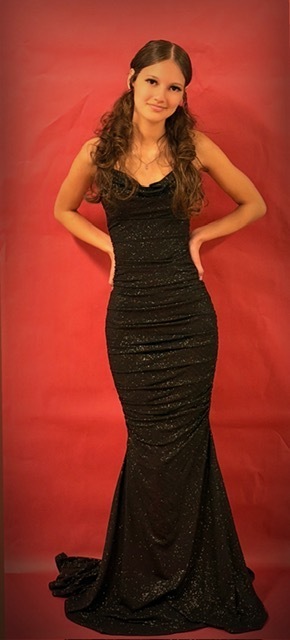 A student poses against a red backdrop wearing a long, form‑fitting black gown with a subtle glittery texture and gentle ruching, standing with hands on hips.