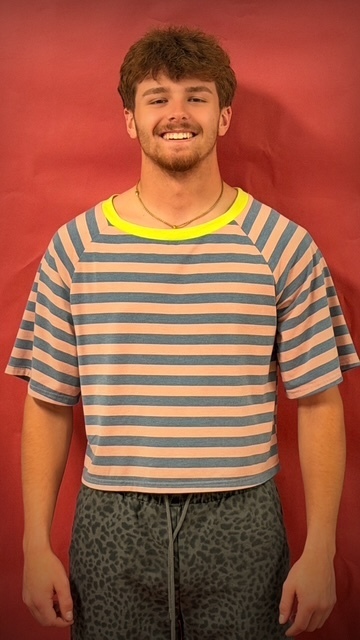 A student stands in front of a red backdrop wearing a cropped striped shirt with a yellow collar and dark patterned shorts.