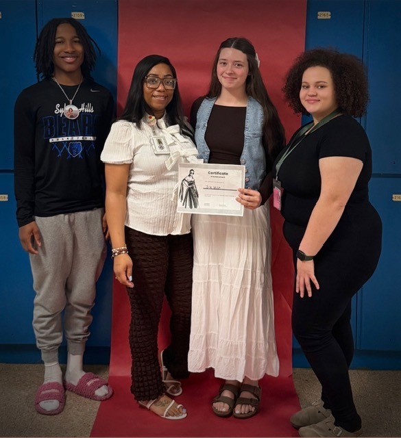 Four students stand in front of blue lockers; one holds a certificate featuring an illustrated historical figure. They pose together to celebrate the recognition.