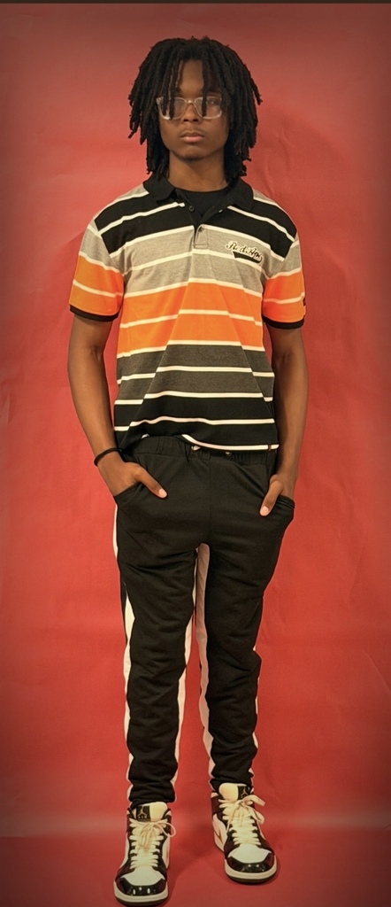 A student poses against a red backdrop wearing a striped polo shirt in orange, gray, white, and black, paired with black pants with white side stripes and multicolored sneakers.