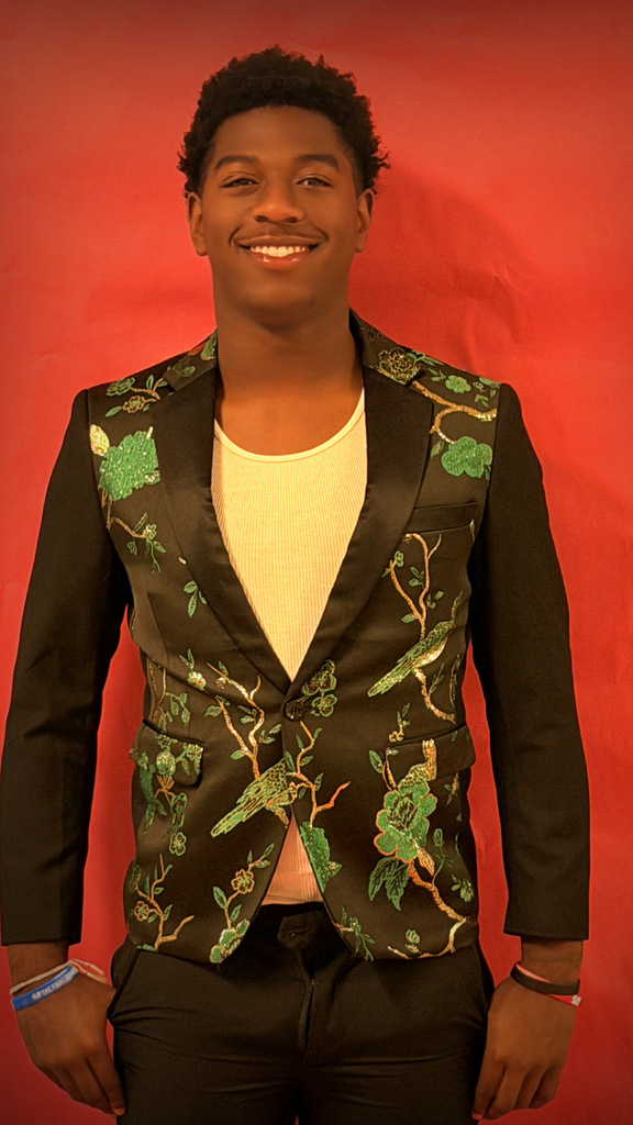 A student poses against a red backdrop wearing a black blazer embroidered with green and gold floral and bird designs over a light ribbed shirt.