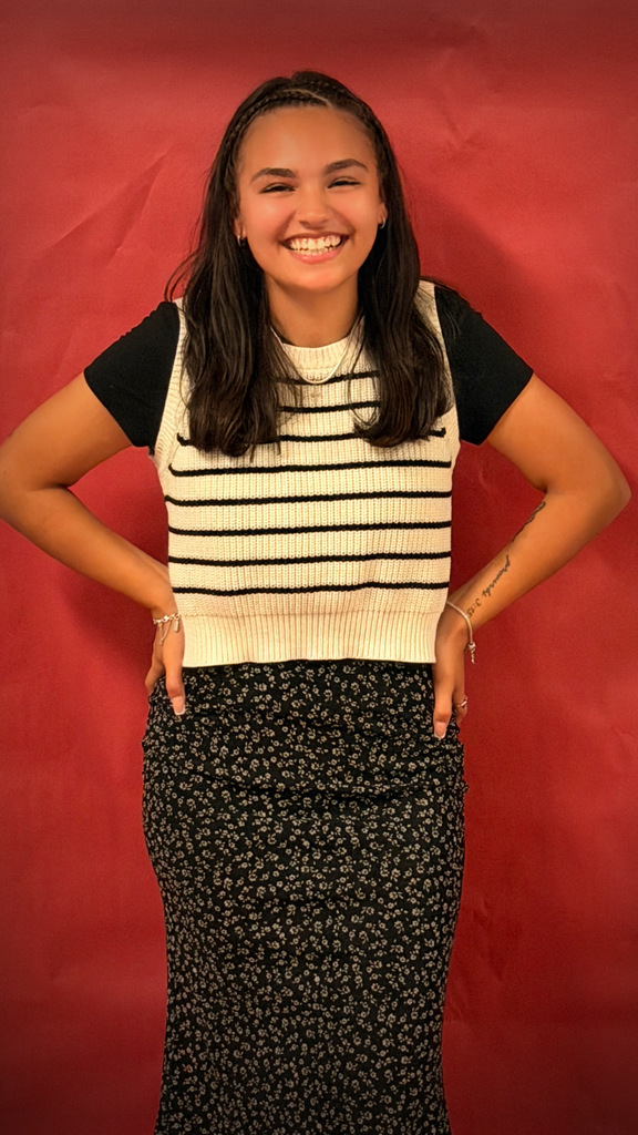 A student poses against a red backdrop wearing a white knitted vest with black stripes over a black shirt and a long black floral skirt, smiling with hands on hips.