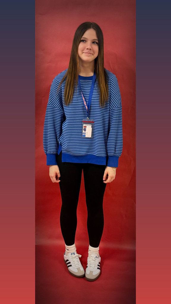 A student stands against a red backdrop wearing a blue and black striped sweater, black leggings, and white sneakers, with a school ID badge on a blue lanyard.