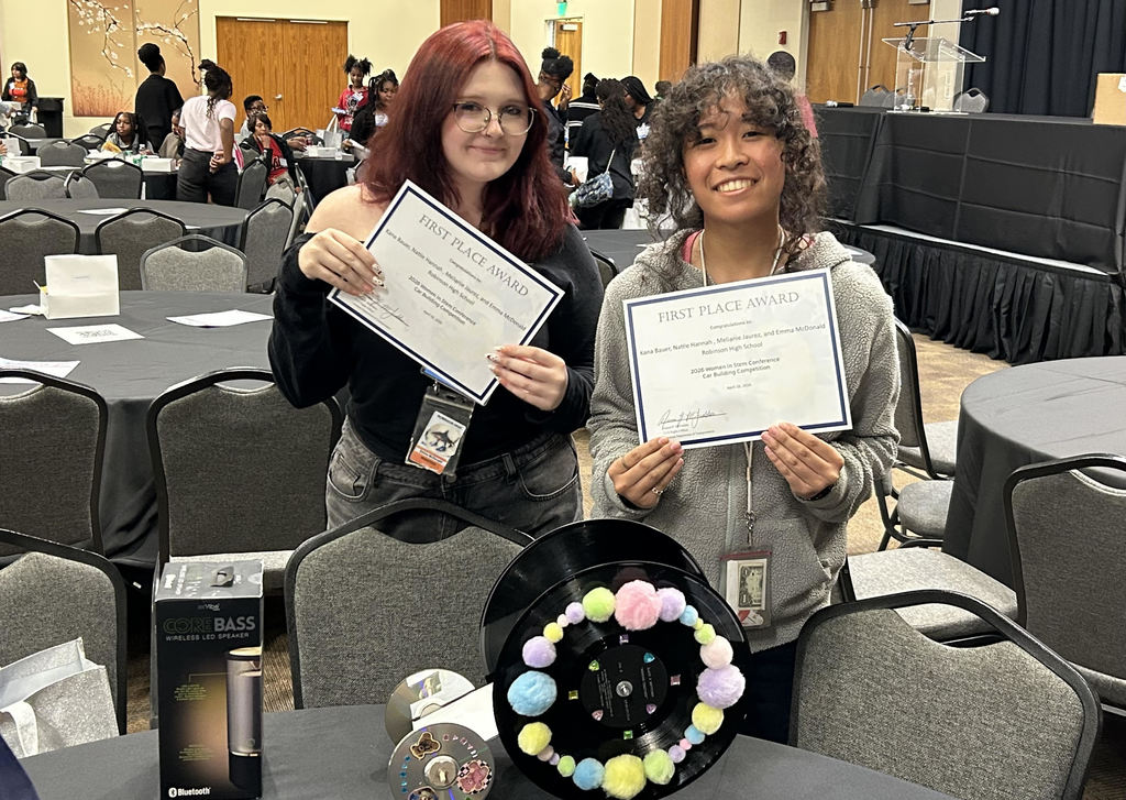 Two Robinson High School students stand in a conference room holding certificates that read “First Place Award” from the 2023 Women in STEM Conference Car Building Competition. A wireless LED speaker prize box and a colorful pom‑pom–decorated project sit on the table in front of them. Other attendees are seated at round tables in the background.