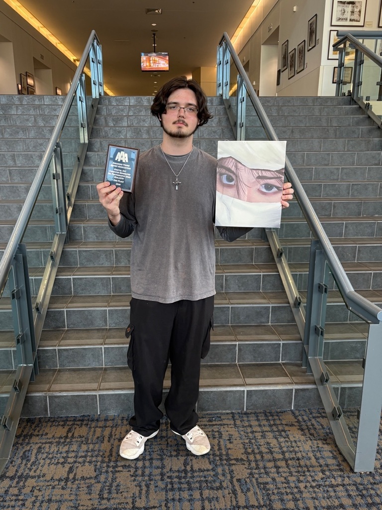 A student stands on a staircase holding a plaque and a framed artwork depicting a close‑up eye partially covered by white fabric. Framed pictures and a television screen appear behind them.