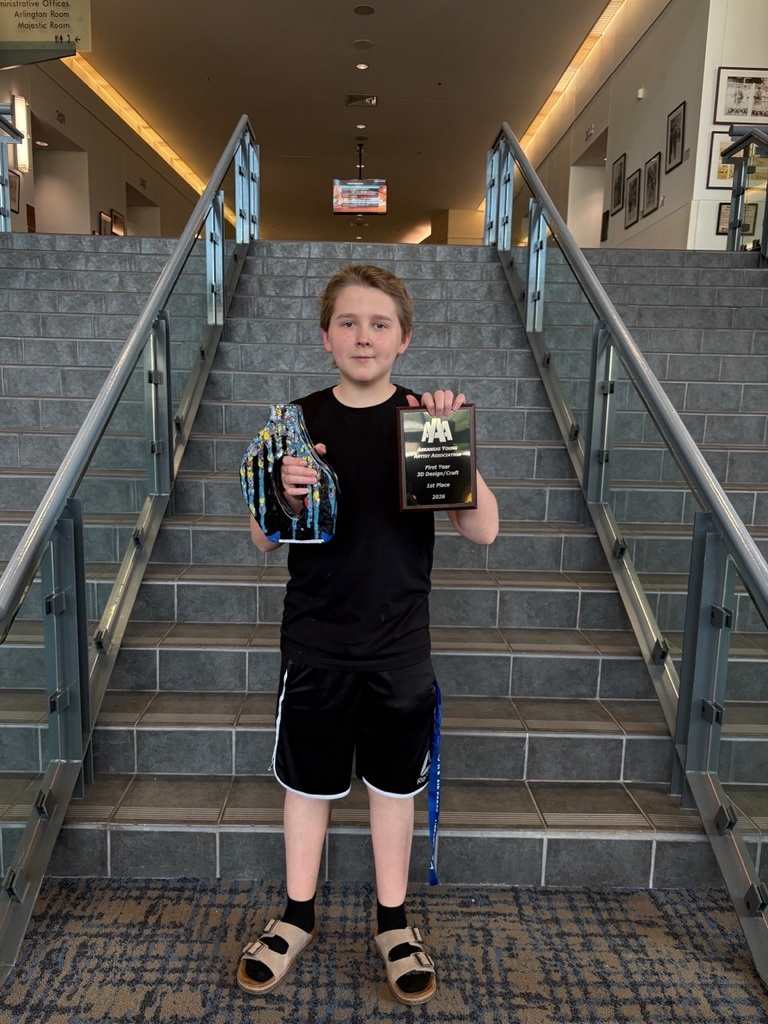 A student stands on a staircase holding a plaque recognizing a 3D Design/Craft award and a decorated art piece. The student is wearing a black shirt, black athletic shorts, black socks, and tan sandals. The background includes tiled steps, metal railings, framed pictures, and directional signage inside the venue.