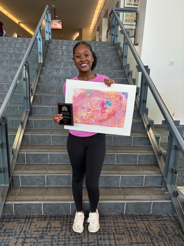 A student stands on a staircase holding an abstract painting with pink, purple, blue, and white textures, along with an award plaque. Framed artwork and a television screen appear in the background.