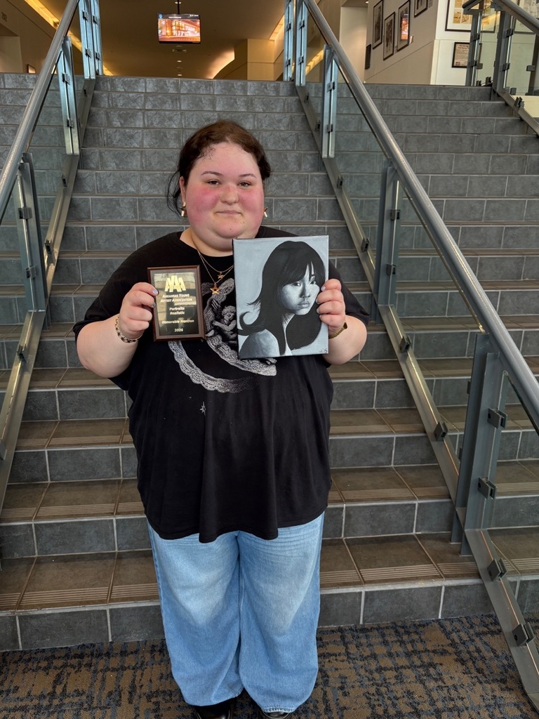 A student stands on a staircase holding a monochrome portrait painting in shades of gray and black, along with an award plaque for painting. The background includes tiled steps and framed artwork.