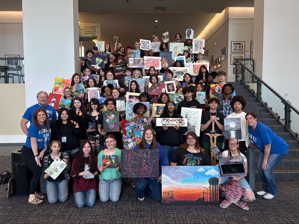 A large group of SHHS art students stand on a staircase holding their artwork, including paintings, drawings, and mixed‑media pieces. Two teachers in blue “Sylvan Hills Bears” shirts stand with them. The group is gathered inside the competition venue with framed artwork and directional signage in the background.