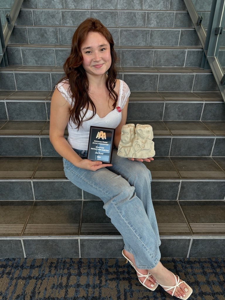 A student sits on a staircase holding a light‑colored sculpture featuring stylized human figures and a plaque recognizing a sculpture award. Glass railings line the staircase.