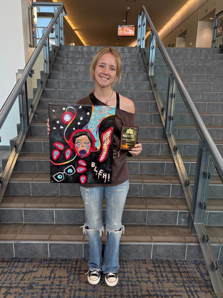 A student stands on a staircase holding a colorful portrait painting with abstract shapes and the word “BLEH!” near the bottom. They also hold a small award plaque. The setting includes tiled steps and glass railings.