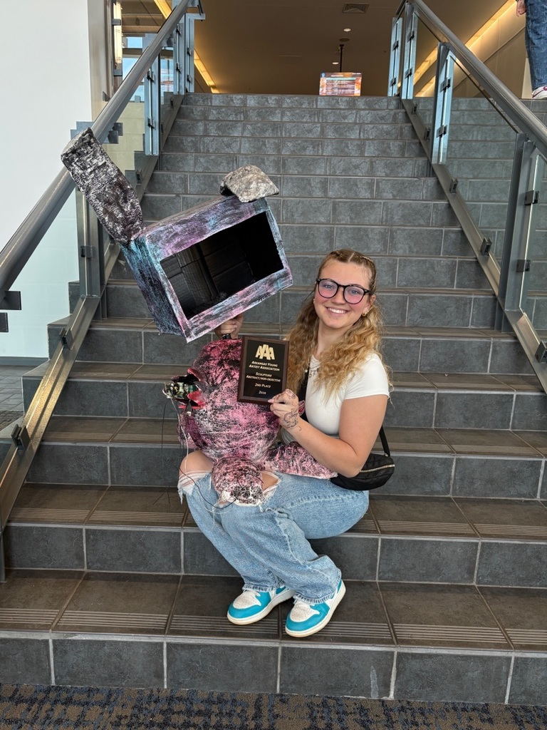 A student sits on a staircase holding a colorful painted box sculpture and a plaque recognizing a presentation award. The sculpture features purple, pink, black, and white designs. The setting includes tiled steps and glass railings.