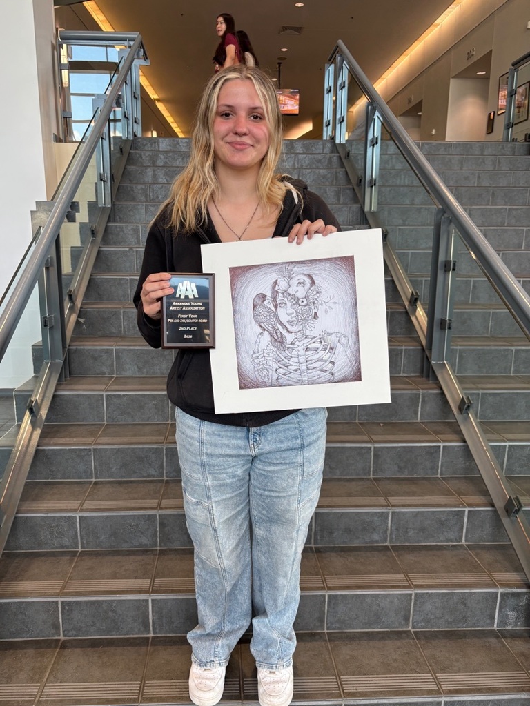 A student stands on a staircase holding a detailed pen‑and‑ink drawing featuring a surreal portrait with multiple faces and a bird. They also hold a plaque recognizing a First Year Pen and Ink award.