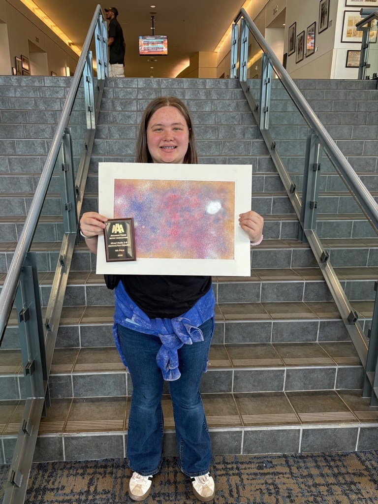A student stands on a staircase holding an abstract painting with pink, purple, blue, and white textures, along with an award plaque. Framed artwork and a television screen appear in the background.