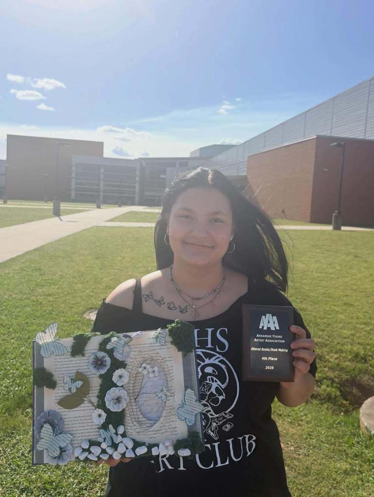 A student stands outside a school building holding an altered‑book artwork decorated with butterflies, greenery, and layered pages. They also hold a plaque for fourth place in altered book/bookmaking at the AYAA competition.