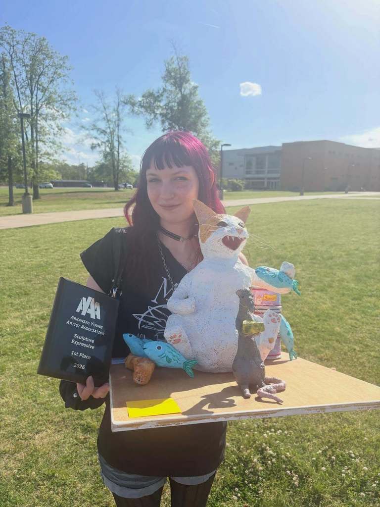 A student stands outside holding a wooden board displaying a colorful sculpture of a white‑and‑orange cat surrounded by blue fish and small sculpted creatures. They also hold a plaque for first place in expressive sculpture at the AYAA competition.