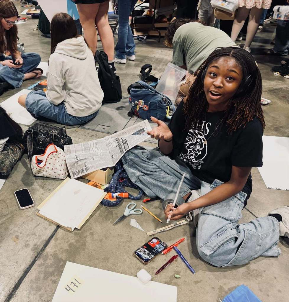 Students sit on the floor of a large indoor space working on on‑site sculpture projects, surrounded by art supplies including scissors, tape, pencils, and paper. Azariah works with a sheet of newspaper as materials are spread around her during the competition.