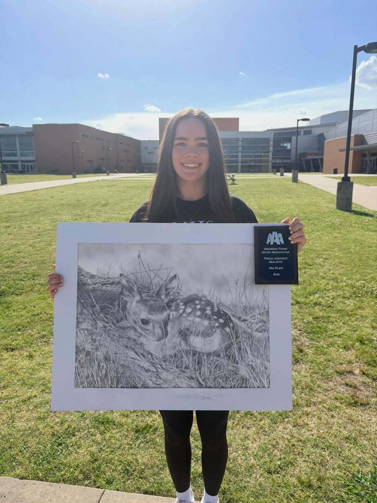 A student stands on a grassy area in front of a school building, holding a detailed pencil drawing of a fawn resting in grass beside a tree. They also hold a plaque for third place in realistic pencil/graphite at the AYAA competition.