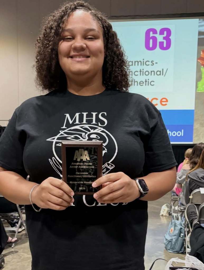 A student stands indoors holding a plaque recognizing an Honorable Mention in the AYAA ceramics functional/utilitarian category. A projected screen behind them displays award information as attendees sit in the background.