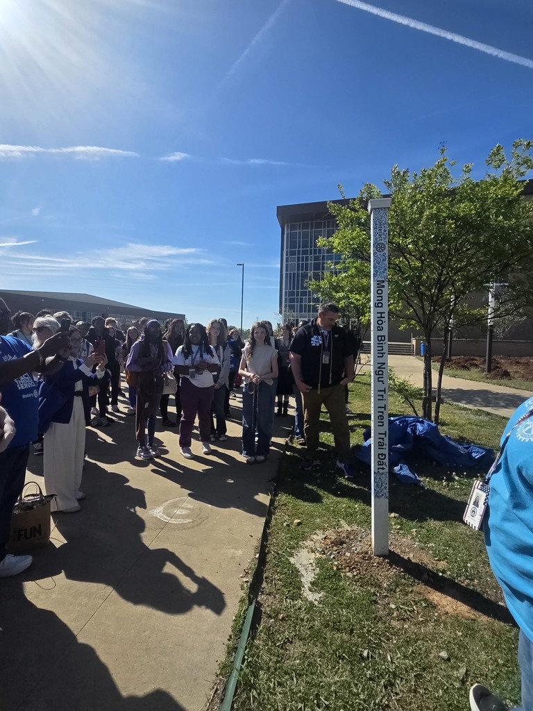 A close-up view of a tall, white, four-sided Peace Pole standing in a mulched garden bed. The phrase 'May Peace Prevail On Earth' is printed vertically down the side."