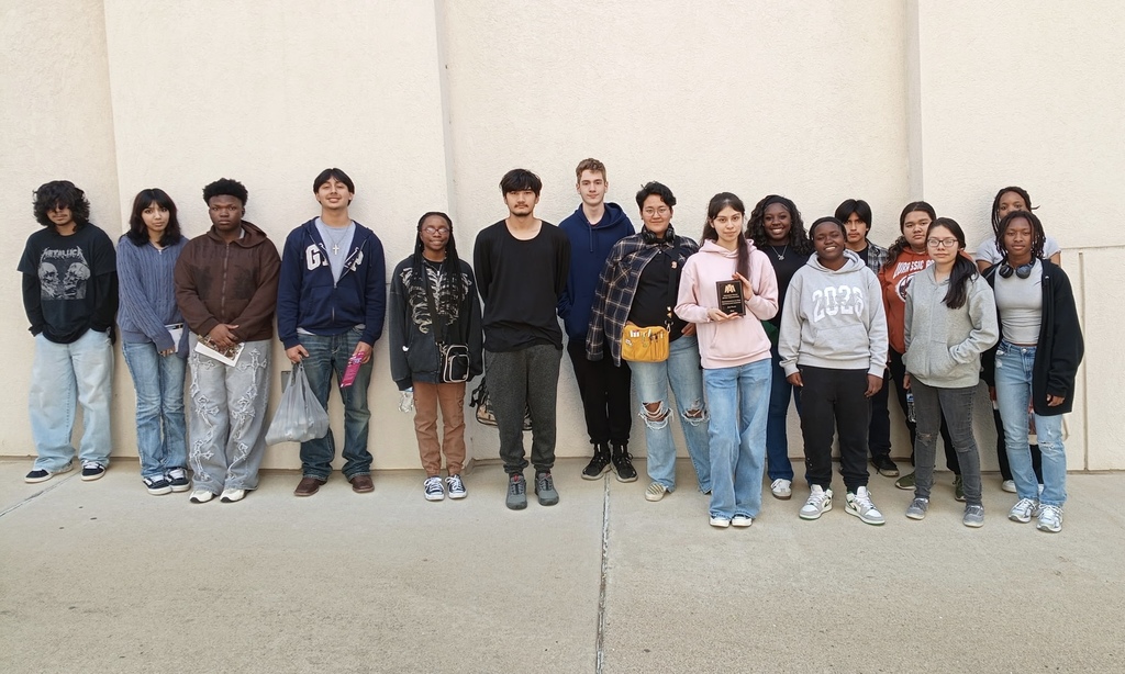 A group of sixteen students stands together outdoors in front of a light-colored wall. One student near the center holds a plaque.
