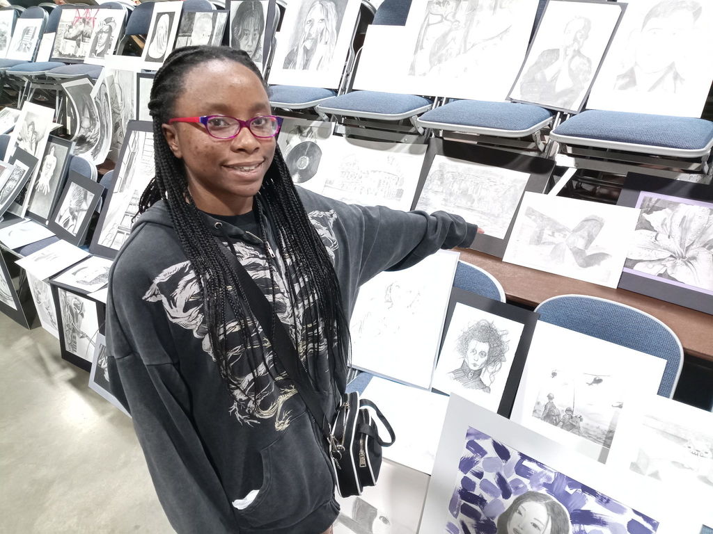 A student wearing glasses and a dark hoodie stands in front of rows of chairs displaying pencil and charcoal drawings. The student points toward one of the artworks.