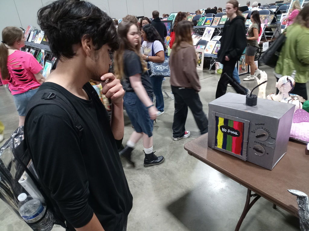 A student stands at a table displaying a gray, sculptural artwork shaped like an old television set with antennas. The screen shows vertical colored bars with the words “NO SIGNAL.” Other artwork and attendees are visible in the background at the AYAA competition.