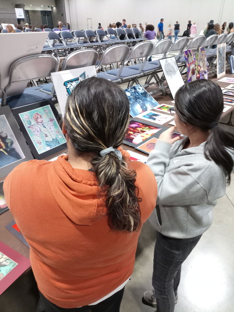 Two people stand at a table covered with colorful drawings and digital artwork. Rows of empty chairs and other attendees are visible in the background of the large event hall.