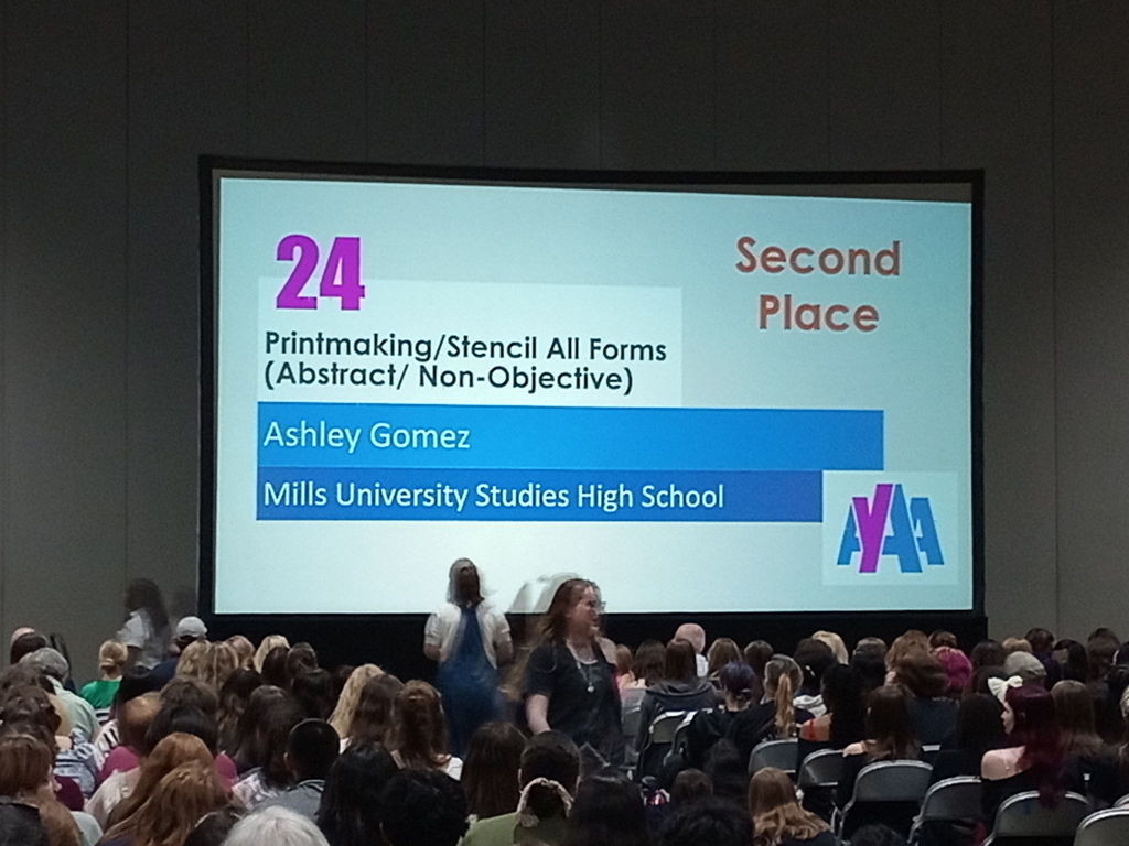 A projection screen at an awards ceremony displays: “Printmaking/Stencil All Forms (Abstract/Non‑Objective) — Ashley Gomez, Mills University Studies High School — Second Place.” Audience members are seated in front of the screen.