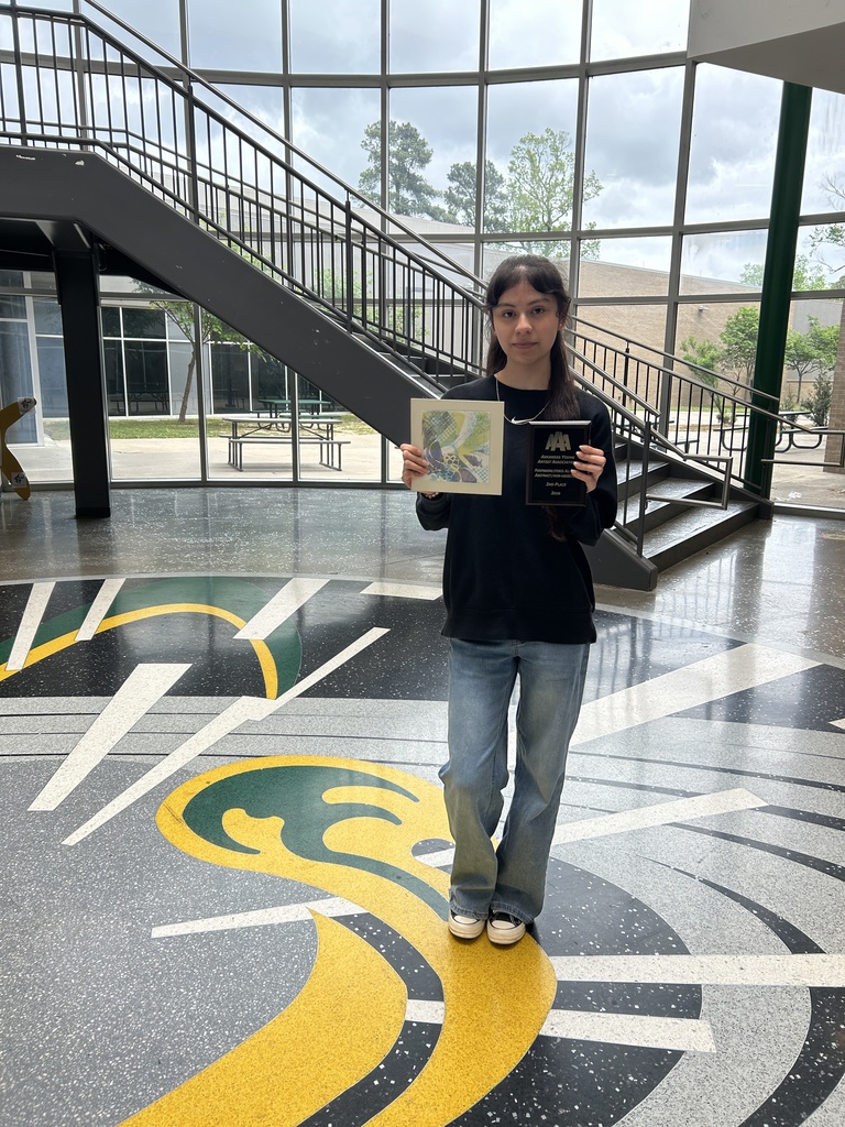 A student stands in a bright lobby holding a plaque and a colorful piece of artwork. Large windows and a staircase are visible in the background.