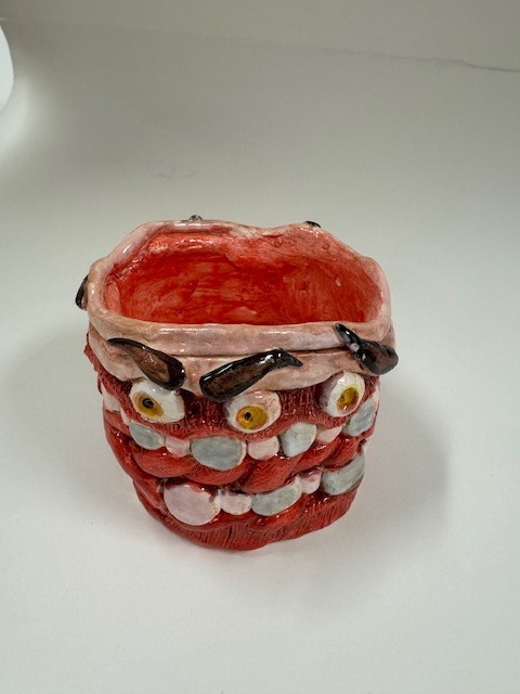 A cylindrical ceramic vessel with a glossy red glaze and raised sculptural features resembling eyes, teeth, and decorative shapes in pink, white, yellow, brown, and blue.