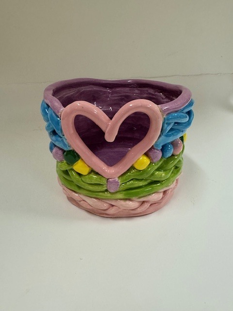 A small ceramic pot with a heart‑shaped opening, made from coiled clay strands glazed in pastel pink, purple, green, and blue with raised yellow and purple dots. The interior is deep purple.