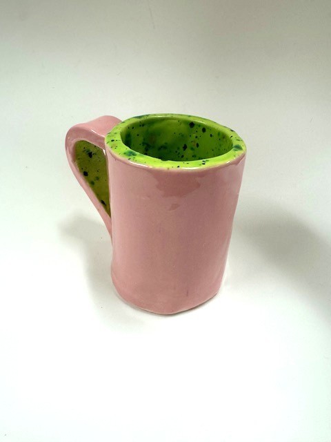 A handmade ceramic mug with a glossy pink exterior and a bright green speckled interior. The handle is also glazed green on the inside.