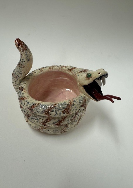 A ceramic bowl formed by a coiled snake with a speckled cream, brown, and gray body. The snake’s head is raised with fangs and a red tongue, and the bowl interior is glossy pink.